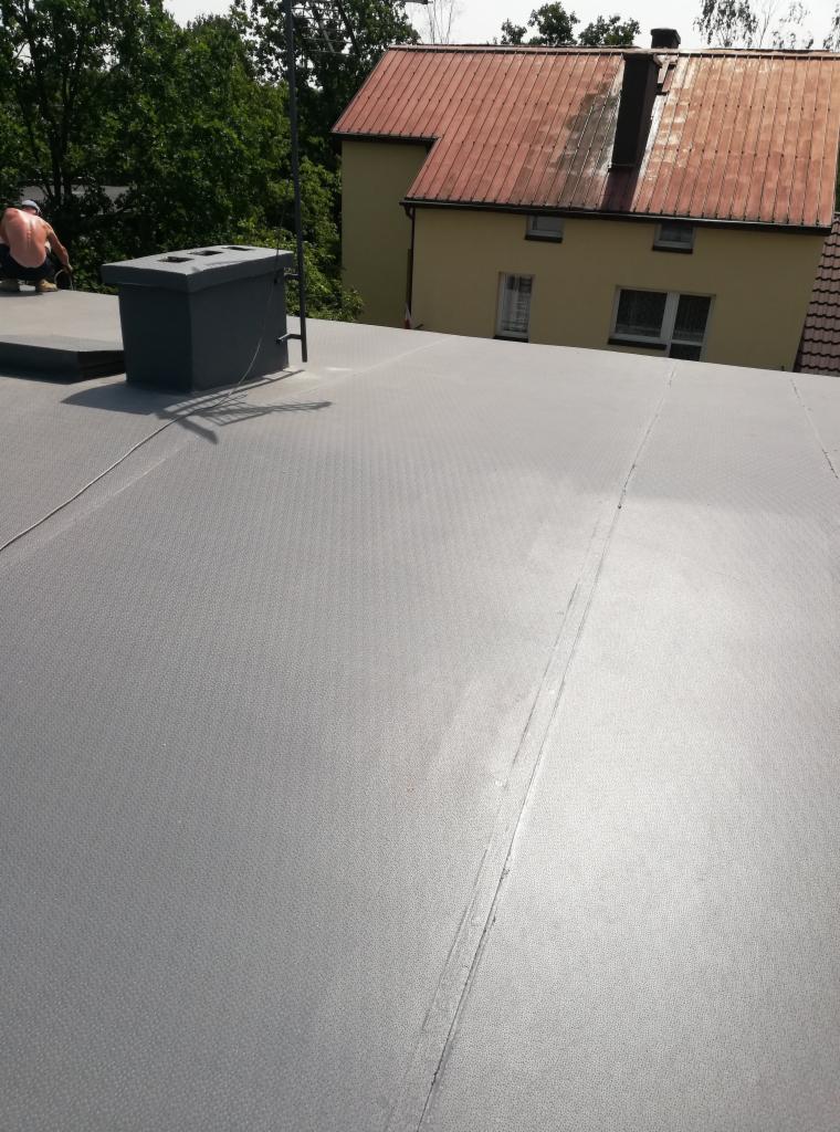 Szary, płaski dach pokryty papą termozgrzewalną z widocznym kominem wentylacyjnym i pracownikiem w oddali, na tle budynku z pomarańczową dachówką.