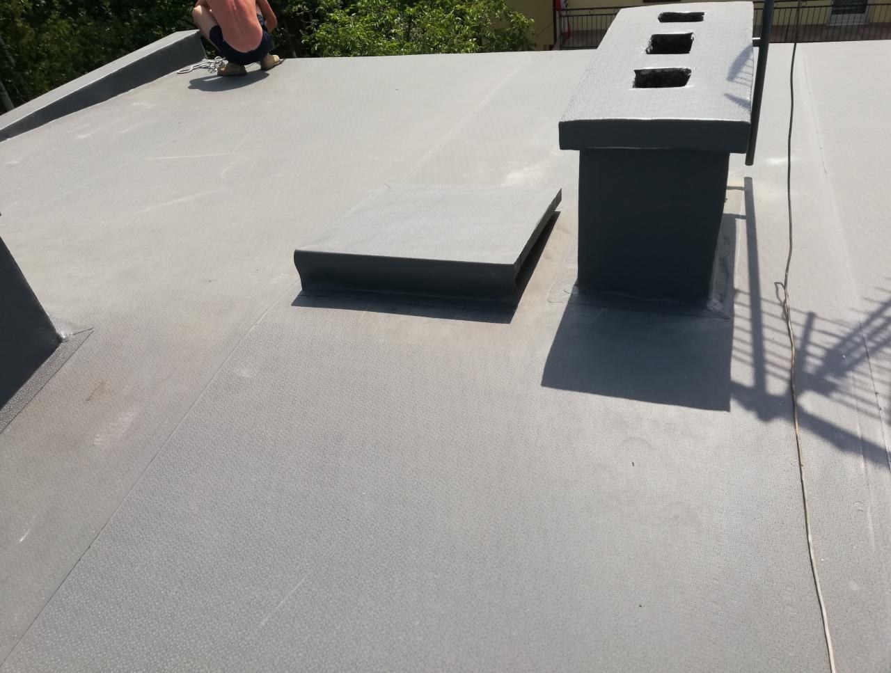 Szary, płaski dach pokryty papą termozgrzewalną z widocznymi kominami i pracownikiem w tle, wykonującym prace dekarskie.