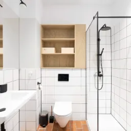 Jasna łazienka z terakotową podłogą, białymi płytkami, kabiną prysznicową z czarną armaturą i drewnianą szafką nad toaletą.