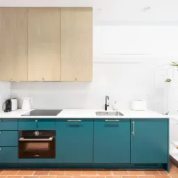 Nowoczesna kuchnia z turkusowymi szafkami, złotymi uchwytami, białym blatem, drewnianymi szafkami wiszącymi i terakotową podłogą, uzupełniona minimalistycznym regałem z roślinami.