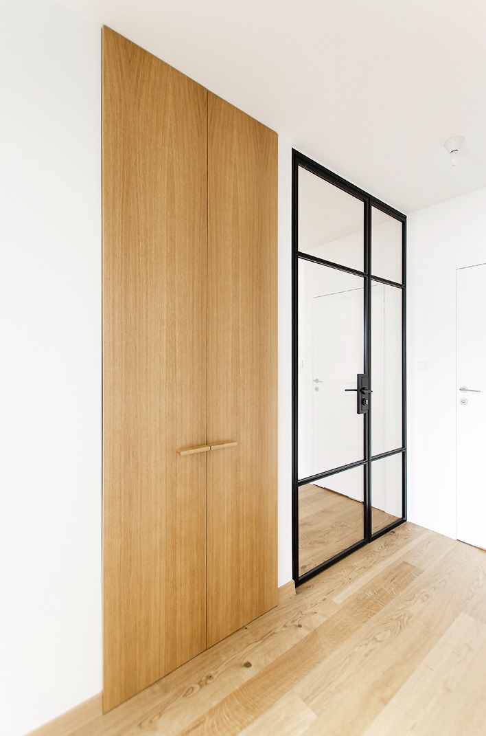 Minimalistyczne wnętrze: wysoka, drewniana szafa w zabudowie, szklane drzwi w czarnej ramie, jasna drewniana podłoga, białe ściany.