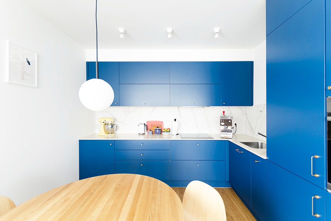 Niebieska kuchnia z białym blatem i marmurowym backsplashiem, okrągły drewniany stół z krzesłami na pierwszym planie, żółty mikser KitchenAid na blacie.