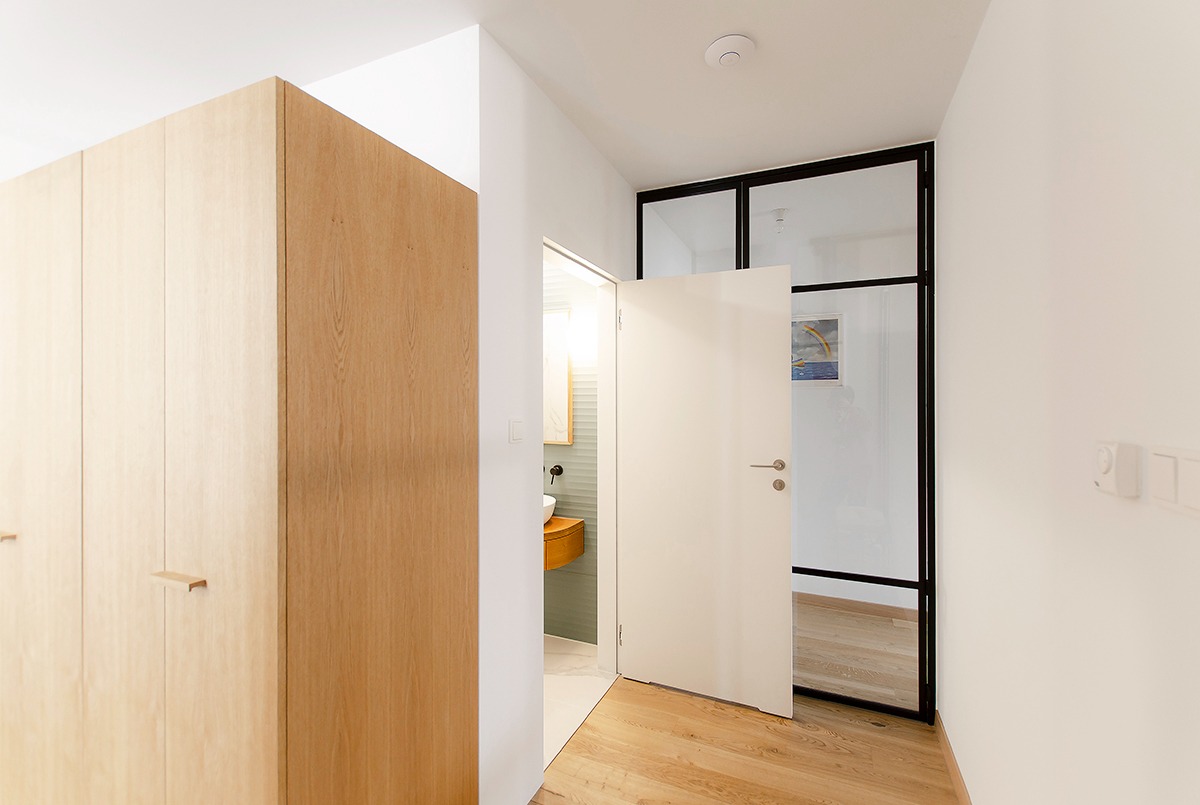 Jasne drewniane szafy w przedpokoju z otwartymi drzwiami do łazienki z okrągłą umywalką i szklaną ścianą z czarnymi ramami.