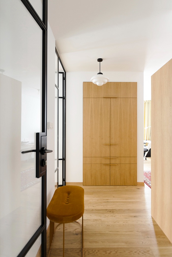 Nowoczesny przedpokój z drewnianą zabudową szafy, szklaną ścianą z czarnymi szprosami i musztardowym pufem na złotych nóżkach.