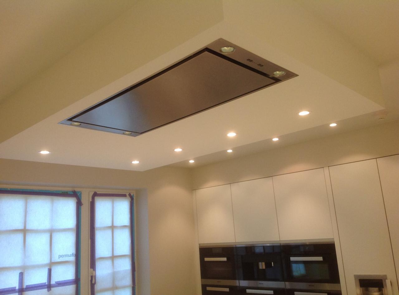 Nowoczesna kuchnia z minimalistyczną zabudową, wpuszczonym okapem ze stali nierdzewnej i oświetleniem punktowym LED, okno zabezpieczone folią malarską.