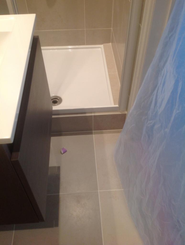 Biały brodzik z odpływem, brązowa szafka pod umywalką i szare płytki podłogowe, zabezpieczone folią malarską, widok z góry.