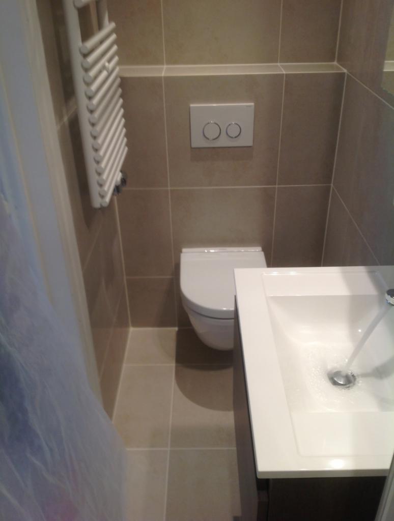 Nowoczesna łazienka z beżowymi płytkami, wiszącą toaletą, grzejnikiem drabinkowym i białą umywalką z baterią, z której leje się woda.