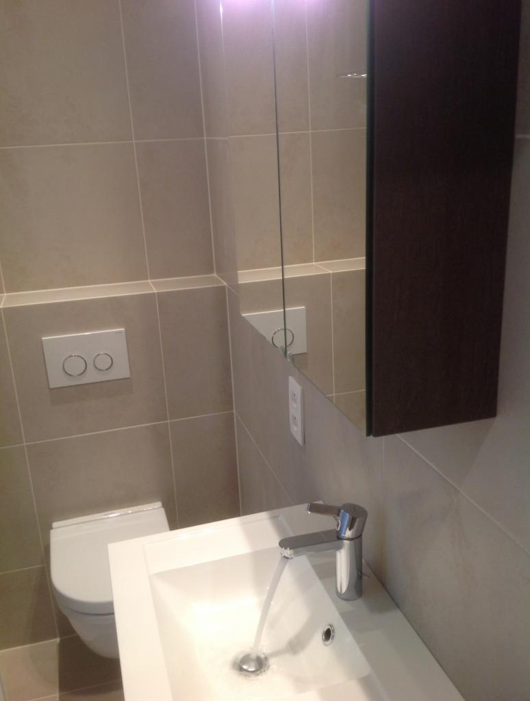 Nowoczesna łazienka z beżowymi płytkami, białą umywalką z chromowaną baterią, spłuczką podtynkową i lustrem z szafką, woda leje się z kranu.