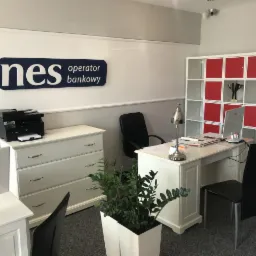 Jasne biuro z biurkiem, komputerem, lampką, regałem z czerwonymi wstawkami, drukarką na komodzie i logo 'Fines operator bankowy' na ścianie.