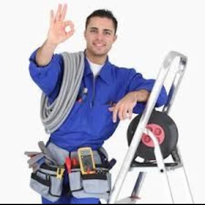 Uśmiechnięty monter w niebieskim kombinezonie, z pasem narzędziowym i zwiniętym przewodem na ramieniu, opiera się o aluminiową drabinę, pokazując gest OK.