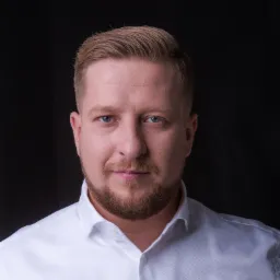 Portret mężczyzny z krótką blond fryzurą i brązową brodą, ubranego w białą koszulę na czarnym tle.