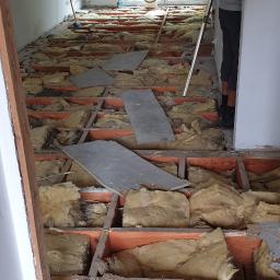 Kad - Demontaż podłogi w mieszkaniu: odsłonięta konstrukcja drewniana z izolacją z wełny mineralnej, widoczny fragment ściany i sylwetka pracownika z poziomicą.