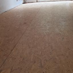 Kad - Przygotowanie podłoża pod podłogę: płyty OSB ułożone na dużej powierzchni w pustym pomieszczeniu z białymi ścianami i widokiem na zielony ogród przez otwarte, przeszklone drzwi balkonowe.