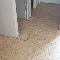 Kad - Montaż podłogi z płyt OSB w trakcie remontu, widoczne płyty przycięte do futryn drzwiowych i ścian działowych z elementami metalowej konstrukcji.