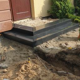 Kad - Granitowe schody wejściowe w trakcie budowy, widoczny wykop, piasek i narzędzia pomiarowe.