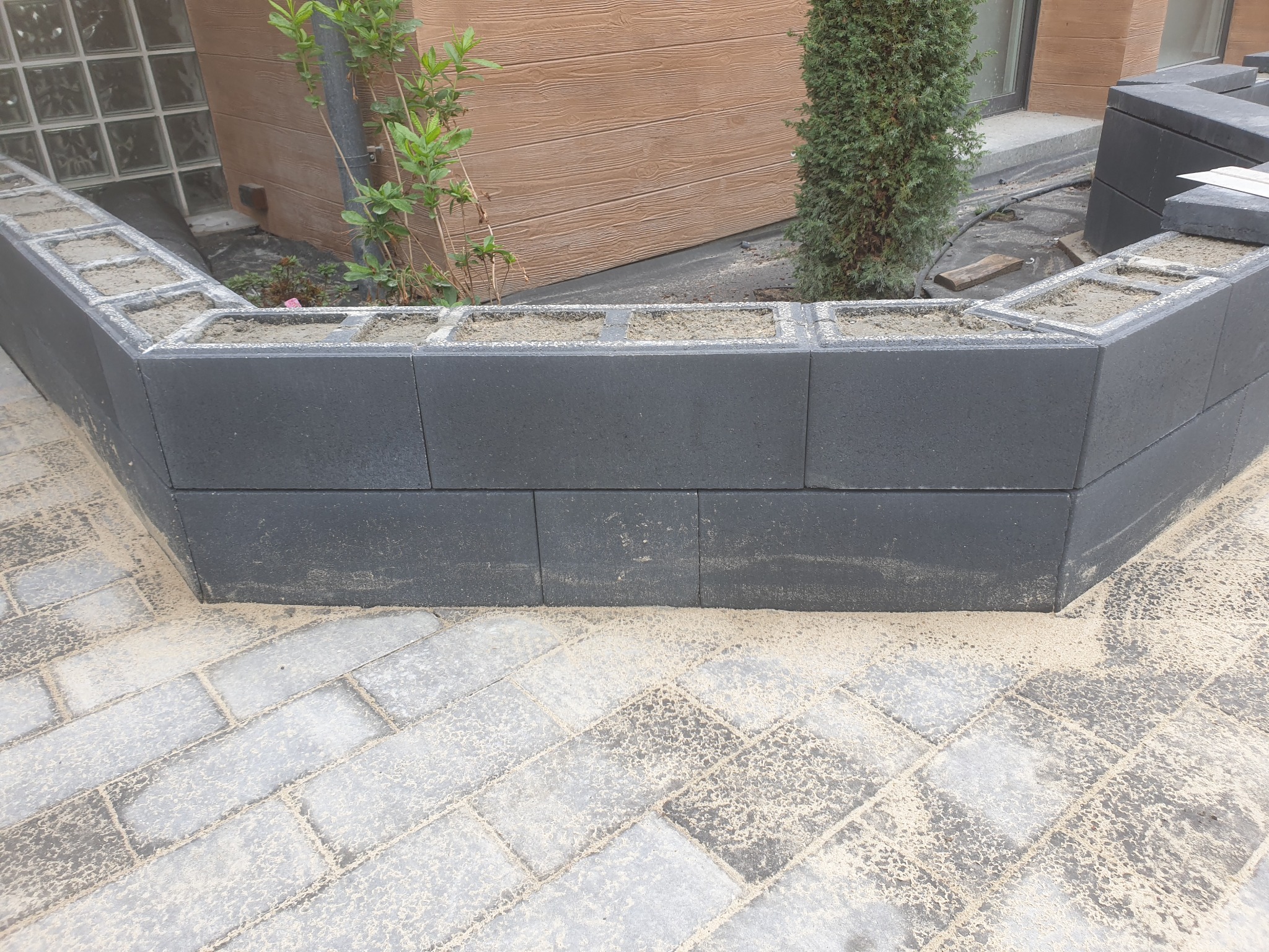 Wzniesiona, ciemna, betonowa obudowa rabaty kwiatowej z pustaków, wypełniona ziemią, z kostką brukową u podstawy, w otoczeniu zieleni i ściany budynku.
