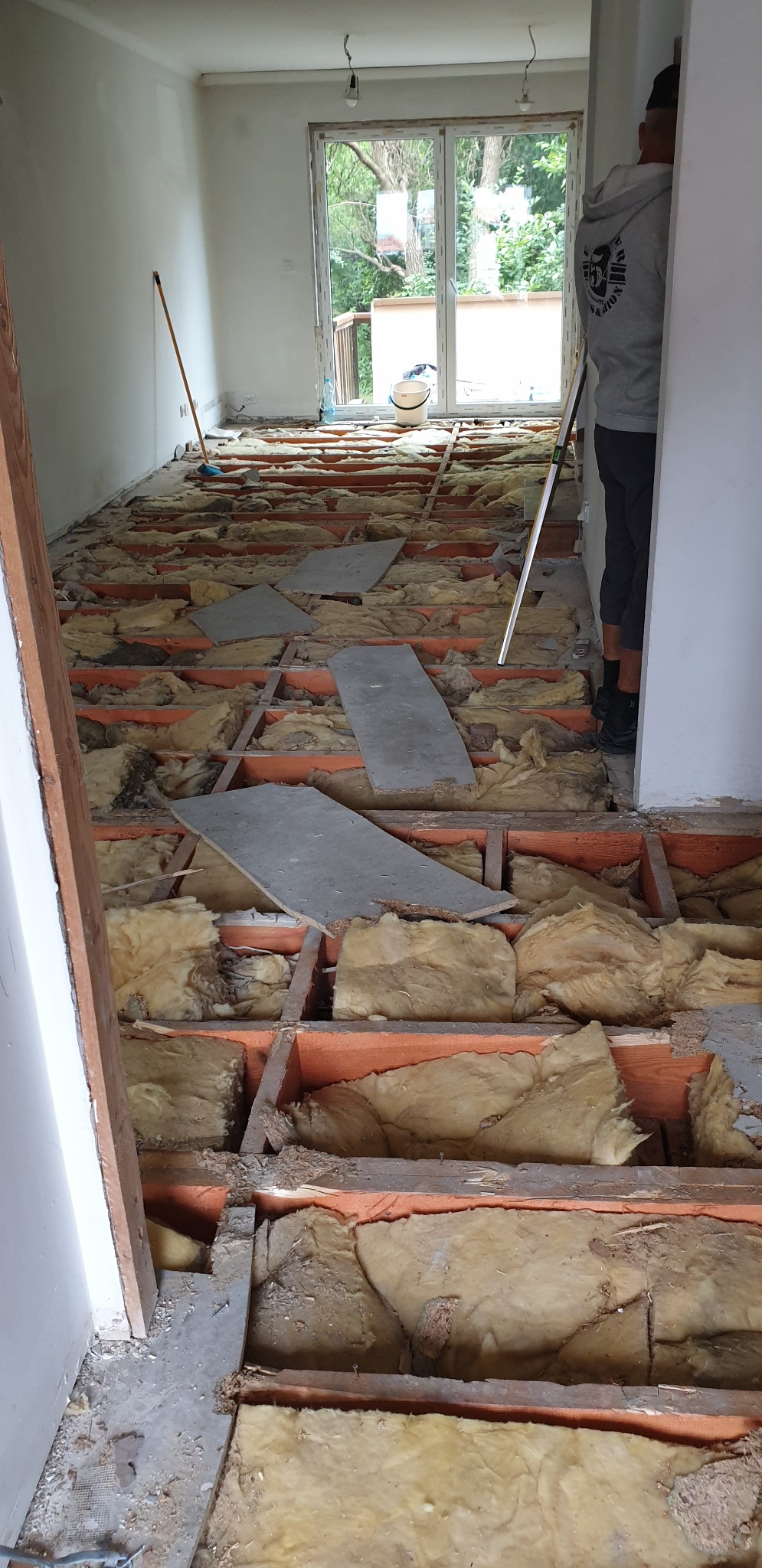 Demontaż podłogi w mieszkaniu: odsłonięta konstrukcja drewniana z izolacją z wełny mineralnej, widoczny fragment ściany i sylwetka pracownika z poziomicą.