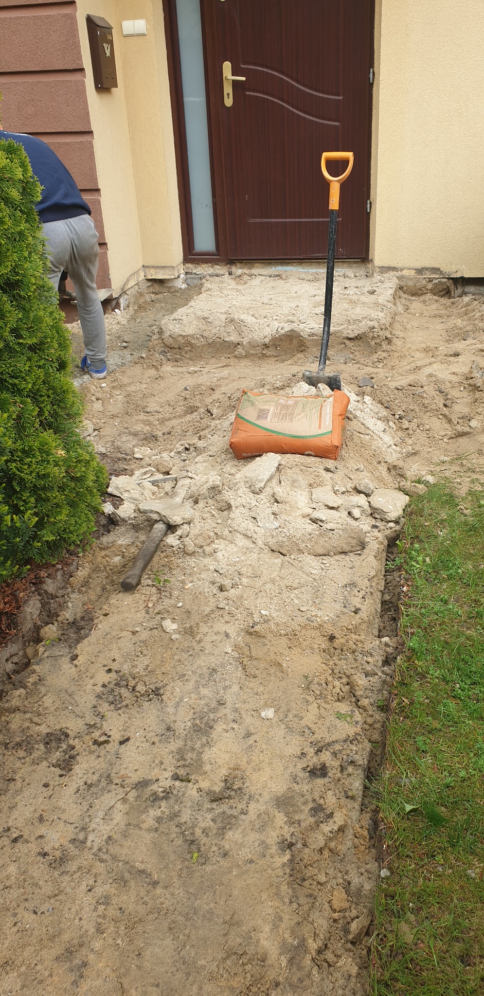 Przygotowanie podłoża przed domem: wykopana ziemia, gruz, łopata, młotek i worek cementu przed wejściem do budynku.