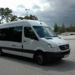 Biały bus Mercedes-Benz Sprinter z ciemnymi szybami i logiem firmy na parkingu z kostki brukowej, w tle drzewa i konstrukcja aquaparku.