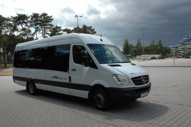 Biały bus Mercedes-Benz Sprinter z ciemnymi szybami i logiem firmy na parkingu z kostki brukowej, w tle drzewa i konstrukcja aquaparku.