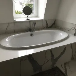 Nowoczesna łazienka z owalną wanną obudowaną białym blatem z konglomeratu i płytkami imitującymi marmur, z małym lusterkiem na parapecie okna.