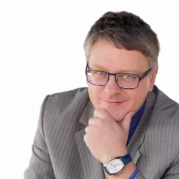 Portret uśmiechniętego mężczyzny w okularach i garniturze w prążki, z zegarkiem na niebieskim pasku, opierającego brodę na dłoni, na białym tle.