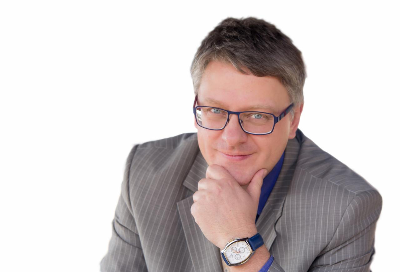Portret uśmiechniętego mężczyzny w okularach i garniturze w prążki, z zegarkiem na niebieskim pasku, opierającego brodę na dłoni, na białym tle.