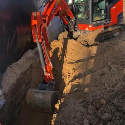Pomarańczowa mini koparka Kubota KX027-4 wykonuje wykop w ziemi, widoczny operator w żółtej kamizelce, obok ściana fundamentowa zabezpieczona czarną folią.