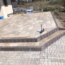 Ułożona jasnoszara kostka brukowa tworząca taras z dwustopniowym wejściem, widoczne narzędzia brukarskie i zapasy kostki w tle.