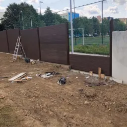 Montaż brązowego ogrodzenia panelowego z kompozytu drewna, widoczna drabina, narzędzia i teren w trakcie prac, w tle boisko sportowe.