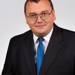 Portret mężczyzny w okularach, czarnym garniturze i błękitnym krawacie, uśmiechający się do kamery na jasnoszarym tle.