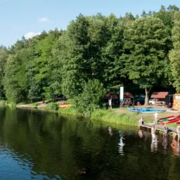 Widok na jezioro z przystanią kajakową, gdzie grupa osób przygotowuje się do spływu, a w tle stoi pomarańczowy autobus i sterta kolorowych kajaków