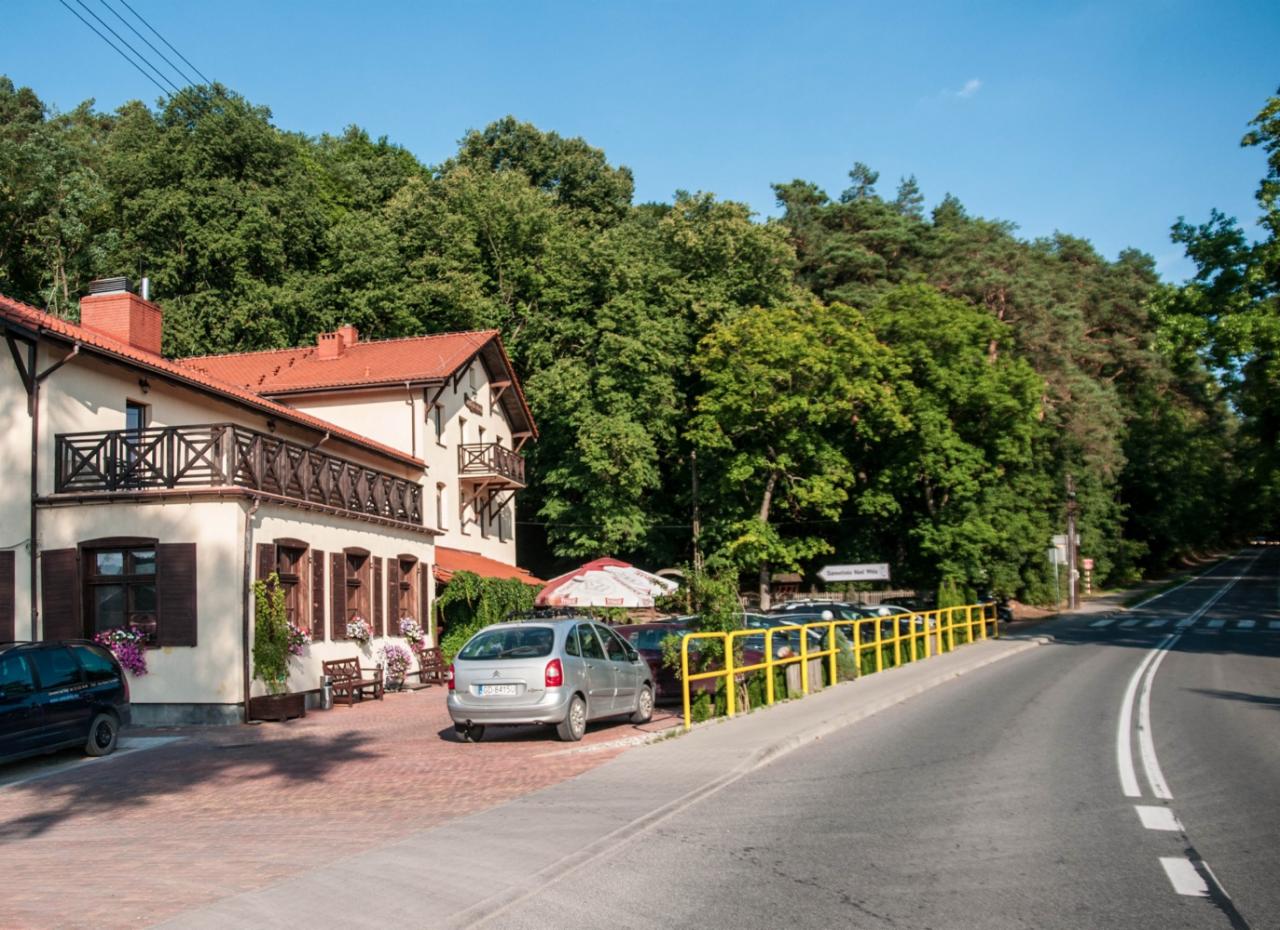 Dwupiętrowy pensjonat z balkonami, czerwonym dachem i brązowymi okiennicami, otoczony zielenią drzew, z zaparkowanymi samochodami i żółtą barierką przy drodze.