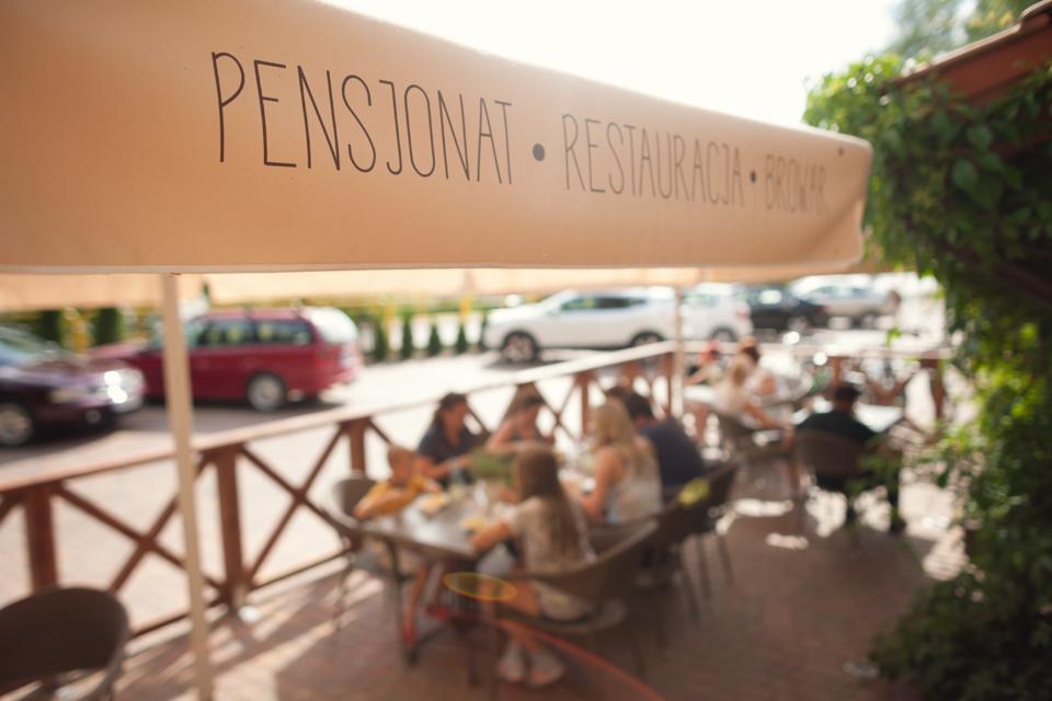 Zadaszony taras pensjonatu z restauracją i browarem, goście siedzą przy stolikach, w tle zaparkowane samochody.
