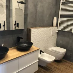 Nowoczesna łazienka z dwoma czarnymi umywalkami nablatowymi, drewnianym blatem, białymi szafkami, szarą płytką ścienną, białymi płytkami 3D, toaletą i bidetem w minimalistycznym stylu.