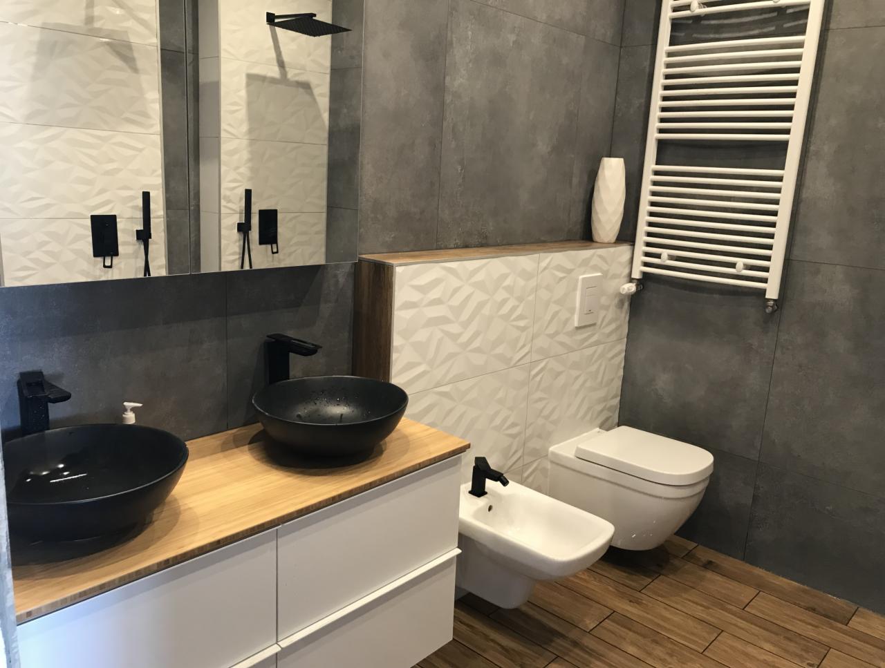 Nowoczesna łazienka z dwoma czarnymi umywalkami nablatowymi, drewnianym blatem, białymi szafkami, szarą płytką ścienną, białymi płytkami 3D, toaletą i bidetem w minimalistycznym stylu.