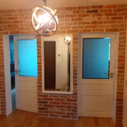 Aranżacja wnętrza z ceglaną ścianą, dwoma białymi, przeszklonymi drzwiami i lustrem w białej ramie, oświetlona lampą w stylu rustykalnym