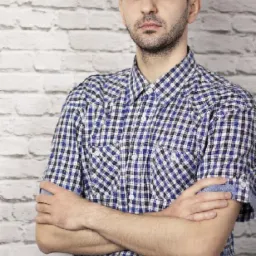 Mężczyzna z brodą, ubrany w niebieską koszulę w kratę, stoi na tle białej ceglanej ściany, z założonymi rękami.