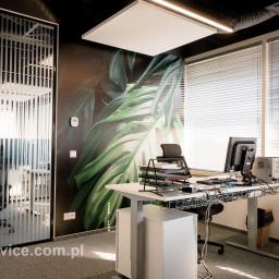 JMP Service Sp. z o. o. - Nowoczesne biuro z biurkiem z regulowaną wysokością, ścianą z motywem roślinnym i szklaną ścianką działową z pionowymi paskami.