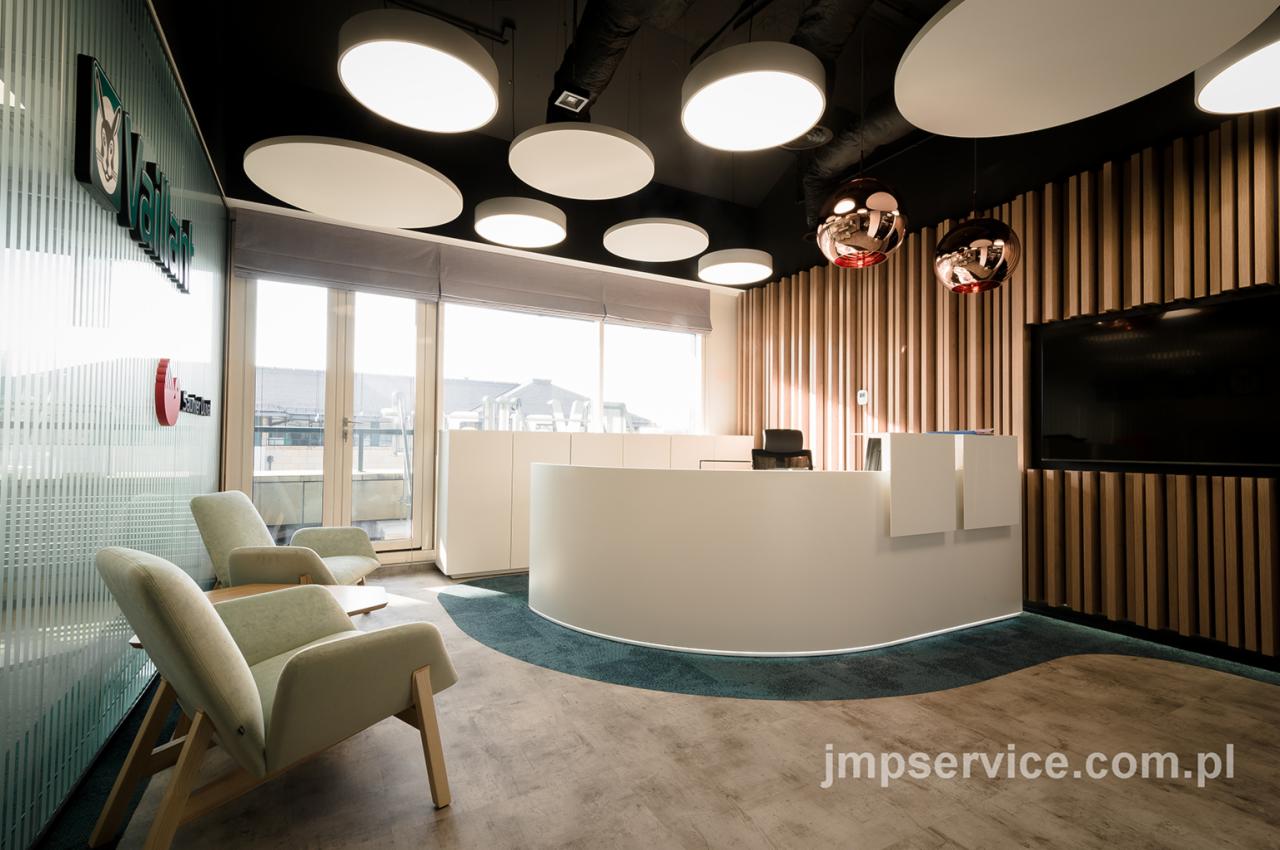 Nowoczesna recepcja biurowa z okrągłą ladą, drewnianą ścianą, wiszącymi lampami w kształcie kul i designerskimi fotelami w strefie oczekiwania.