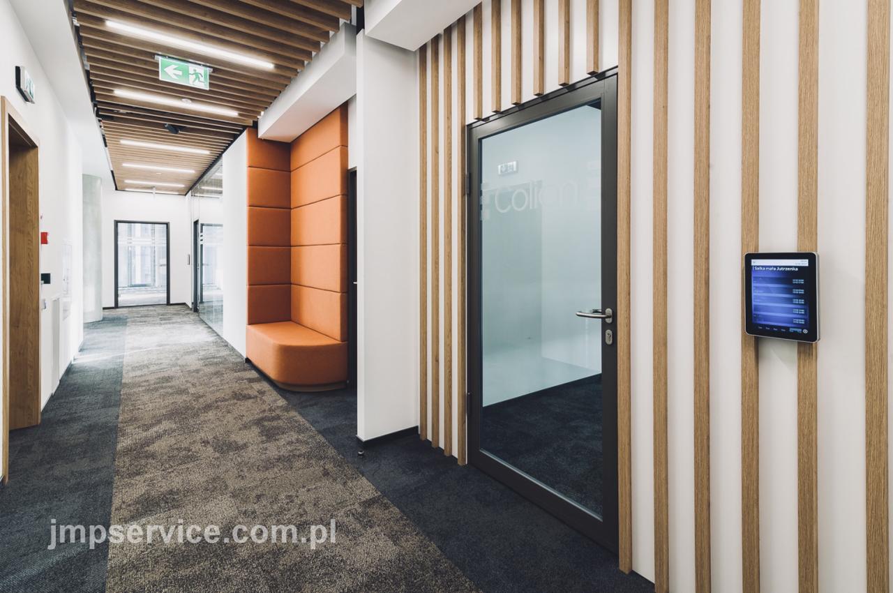 Korytarz biurowy z pomarańczową wnęką do siedzenia, ścianą z drewnianych listew i szklanymi drzwiami z logo firmy, tablet z harmonogramem spotkań na ścianie.