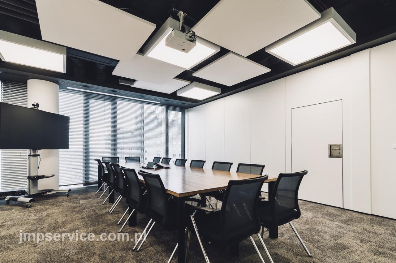 Nowoczesna sala konferencyjna z dużym drewnianym stołem, czarnymi krzesłami, projektorem sufitowym i akustycznymi panelami podwieszonymi do sufitu.