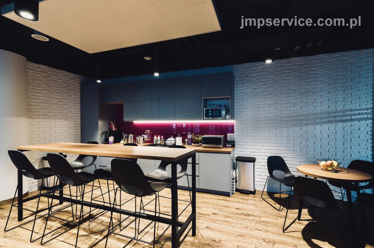 Kącik kuchenny w biurze z długim stołem barowym, krzesłami hokerami, szarymi szafkami i ścianami z białej cegły, oświetlony lampami sufitowymi i podświetleniem LED nad blatem.
