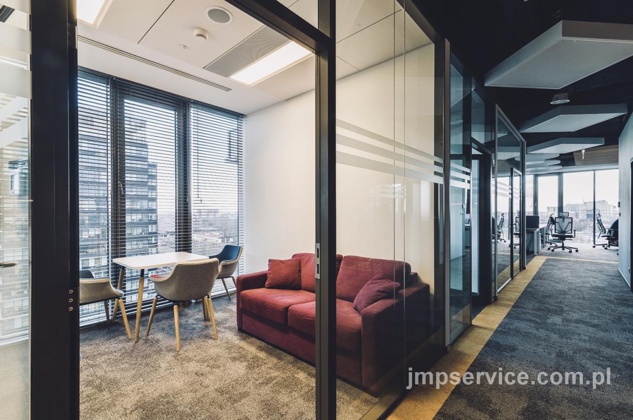 Nowoczesna przestrzeń biurowa z przeszklonymi ścianami, bordową sofą, stolikiem i krzesłami na tle okna z widokiem na miasto.