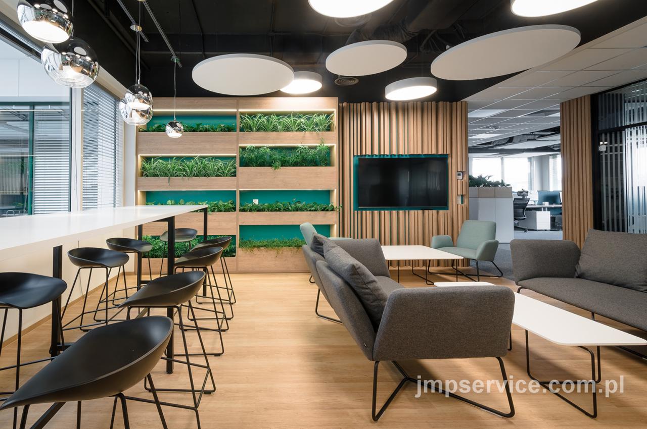 Nowoczesna aranżacja strefy relaksu w biurze z drewnianą ścianą z wbudowanymi roślinami, oświetleniem sufitowym w kształcie dysków, wysokimi krzesłami barowymi i szarymi sofami.