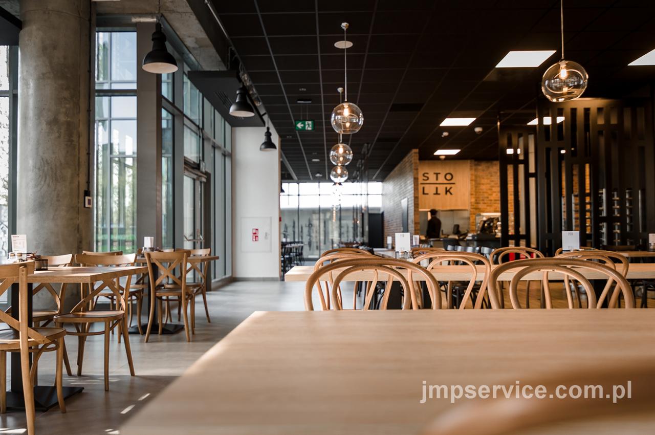 Jasne, przestronne wnętrze restauracji z drewnianymi stołami i krzesłami, betonowymi kolumnami i szklanymi lampami wiszącymi w rzędzie nad stołami, widok na bar w tle.