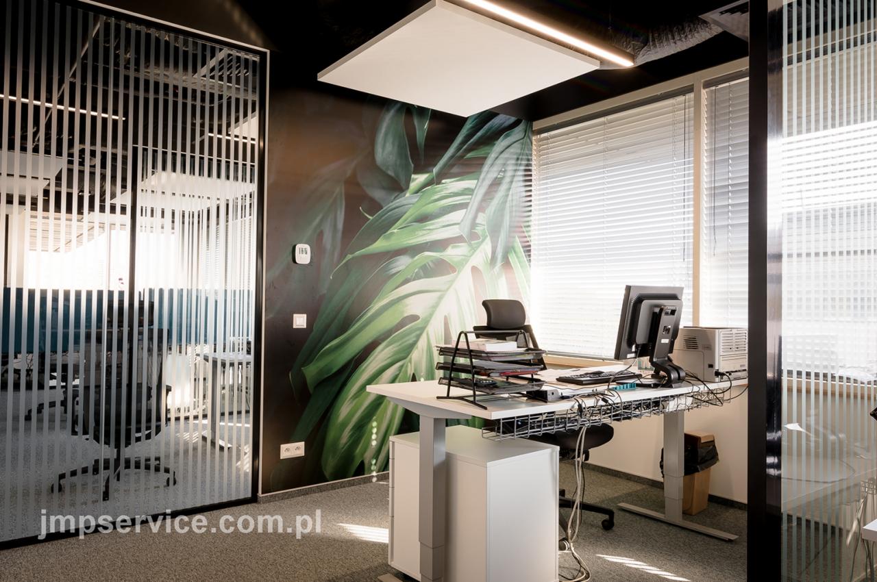 Nowoczesne biuro z biurkiem z regulowaną wysokością, ścianą z motywem roślinnym i szklaną ścianką działową z pionowymi paskami.