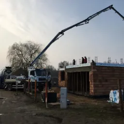 Wylewanie betonu na ściany domu z czerwonej cegły przy użyciu pompy do betonu, widoczne dwie ciężarówki z betonem oraz grupa pracowników na budowie.