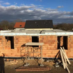 Wznoszenie ścian domu z czerwonej cegły ceramicznej Porotherm, widoczne otwory okienne i drzwiowe, częściowo wykonany wieniec, w tle inne budynki i błękitne niebo z chmurami.