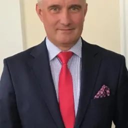 Portret mężczyzny w garniturze z czerwoną krawatem i wzorzystą poszetką, uśmiechający się do kamery na jasnym tle. Elegancki wygląd.